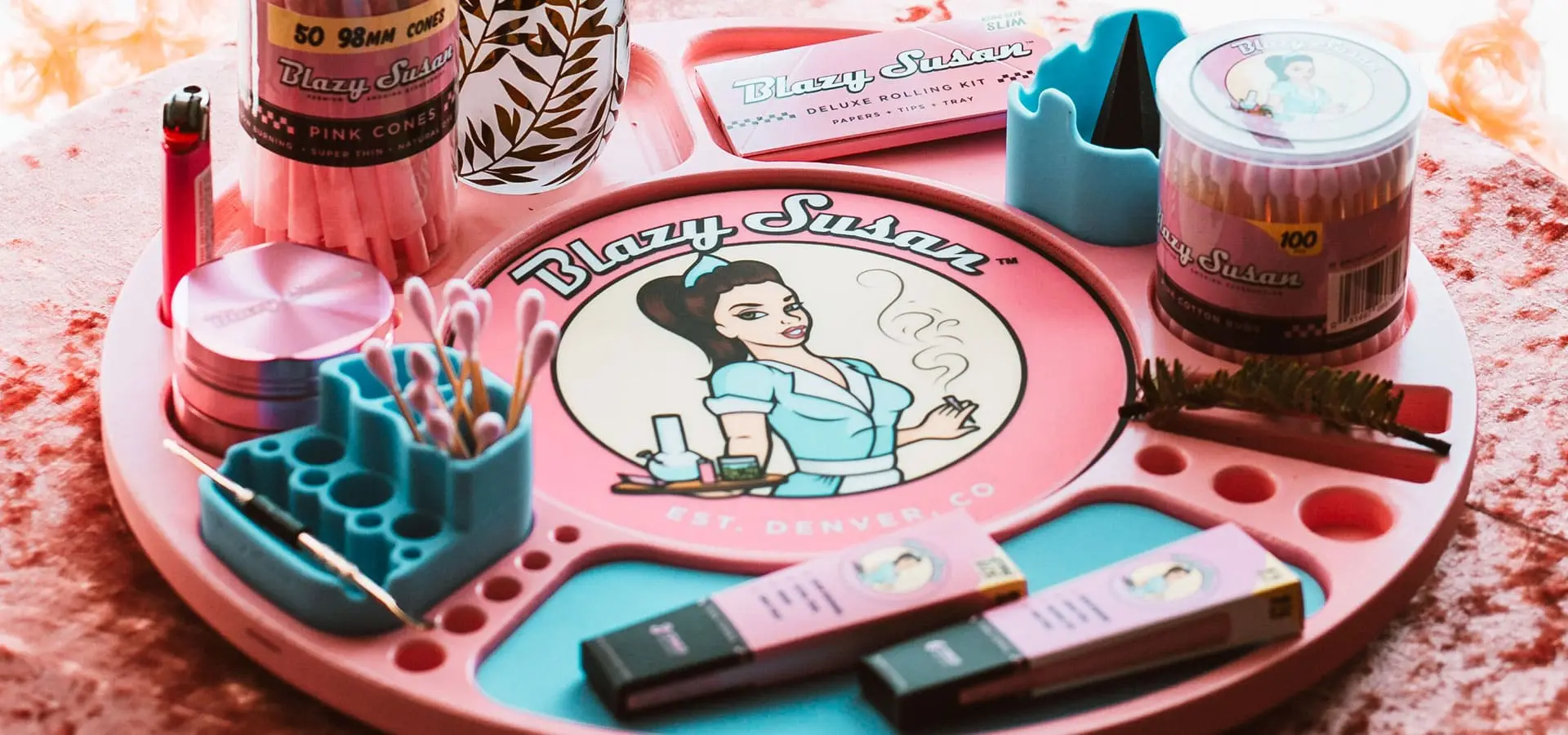 A pink Blazy Susan rolling tray holding cones, papers, a grinder, and cotton buds