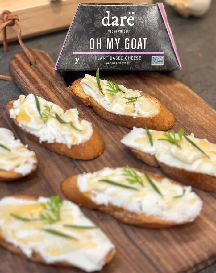 Crostini topped with Darë plant-based cheese and rosemary on a wooden board, with an Oh My Goat box behind
