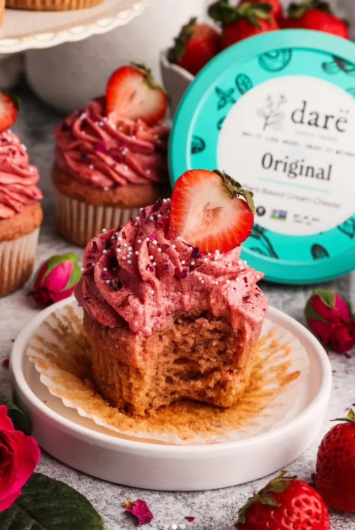 A strawberry cupcake with a bite taken out, in front of a teal Darë Original cream cheese tub