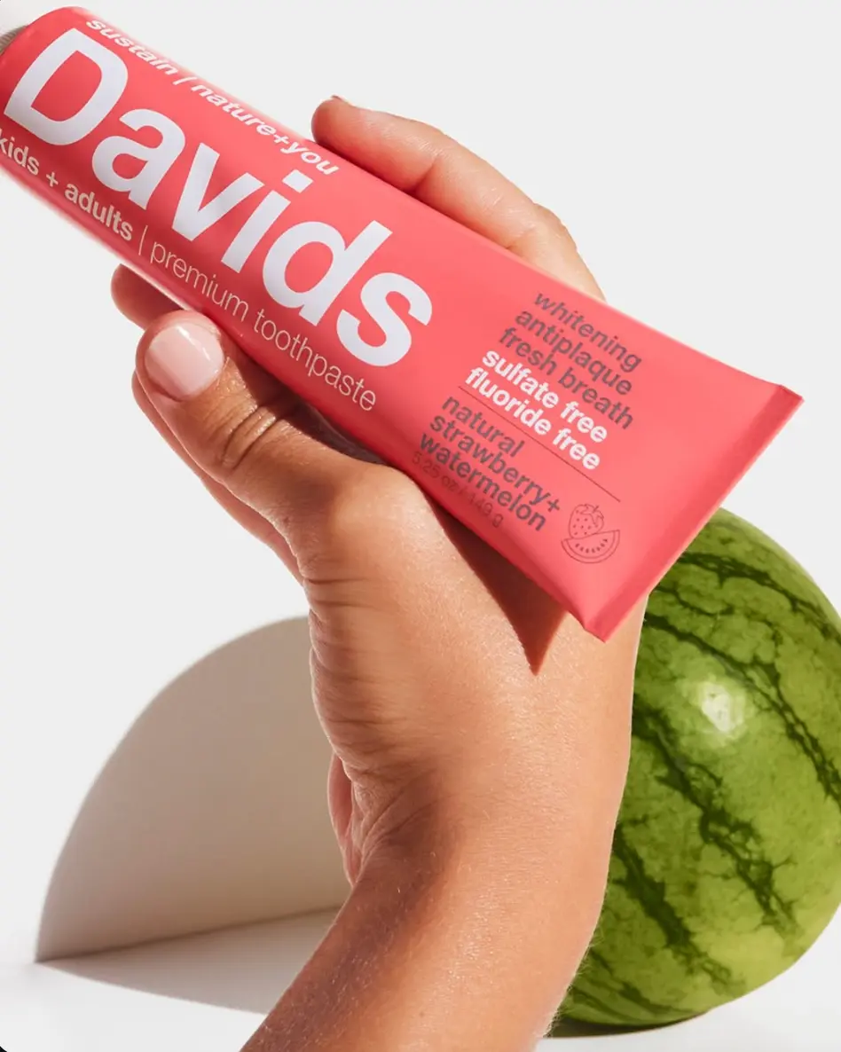A hand holding a Davids strawberry and watermelon toothpaste tube beside a watermelon