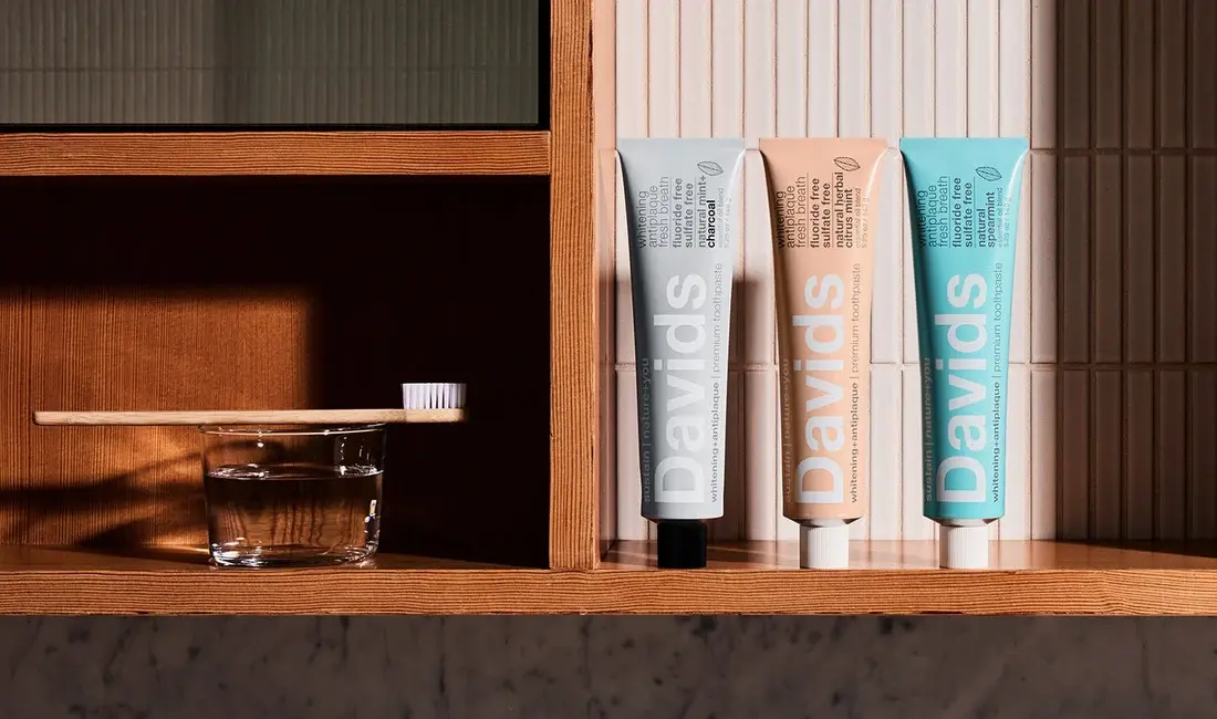 Three Davids toothpaste tubes standing on a wooden bathroom shelf beside a toothbrush