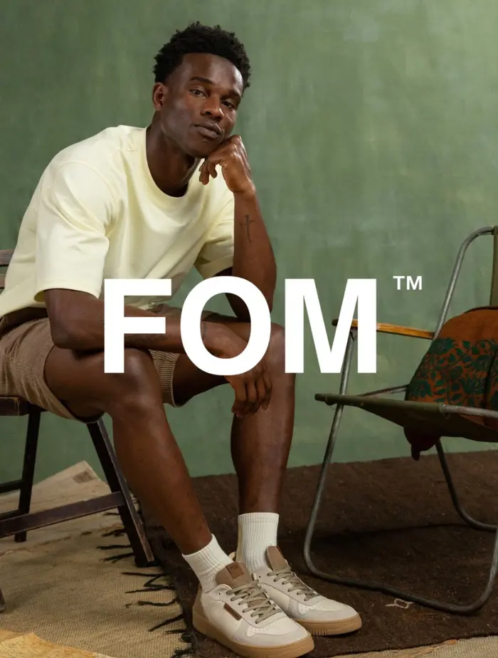 A man seated in a cream tee and shorts, wearing FOM sneakers against a green wall