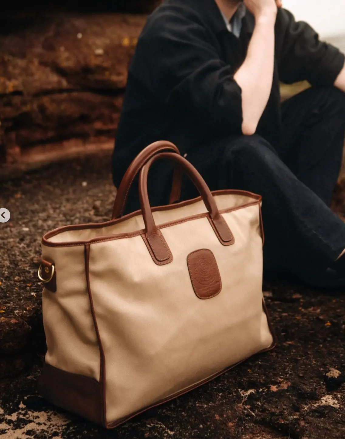 A beige canvas and brown leather Ghurka tote bag on a rock, with a seated person behind it