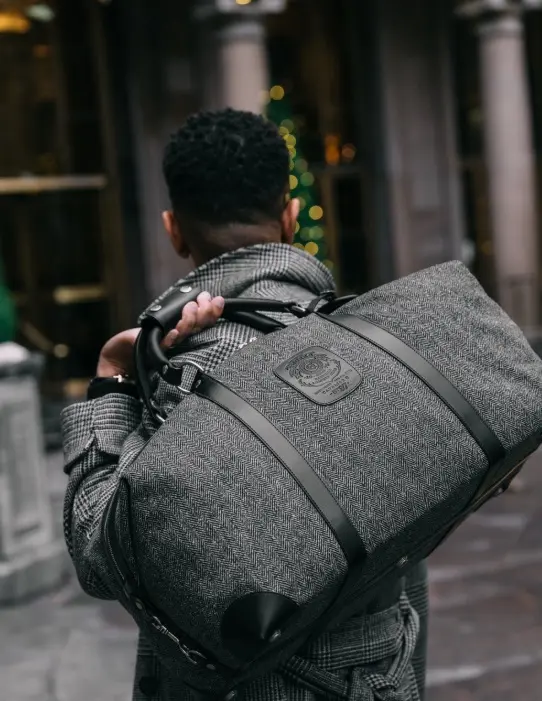 A man carrying a gray herringbone Ghurka duffel bag on his shoulder