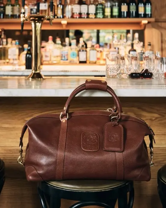 A brown Ghurka leather duffel bag on a bar stool in front of a shelf of bottles