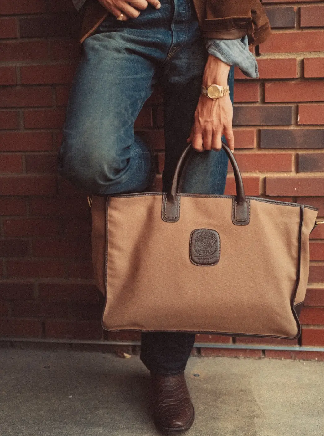 A man leaning against a brick wall, one boot up, holding a tan canvas and brown leather Ghurka tote