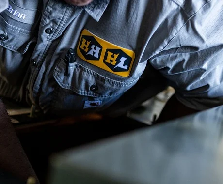 Close-up of a work shirt with the HAHL patch, a technician leaning over equipment