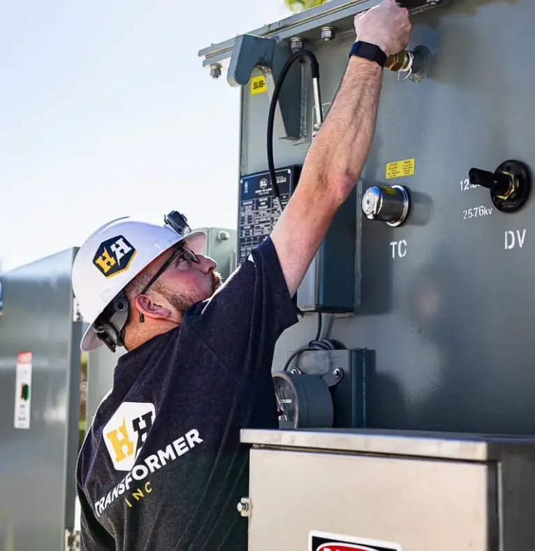 A High to Low Voltage technician in a hard hat reaching up to a transformer control