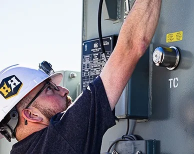 A technician in a hard hat reaching up to adjust the controls on a large electrical transformer
