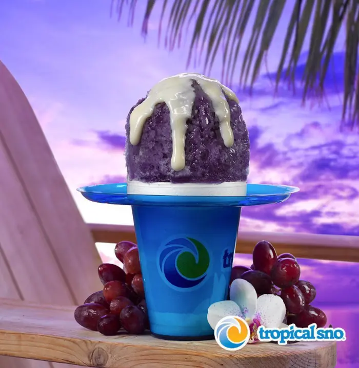 A Tropical Sno cup of grape shaved ice with cream on a blue tray, with grapes and an orchid on a wooden chair