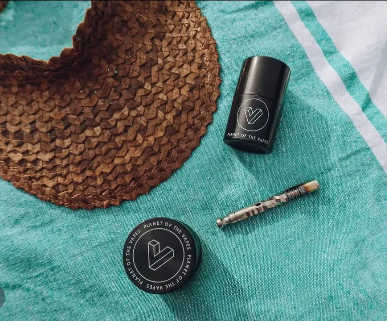 A Planet of the Vapes grinder, jar and glass piece on a teal towel beside a straw hat
