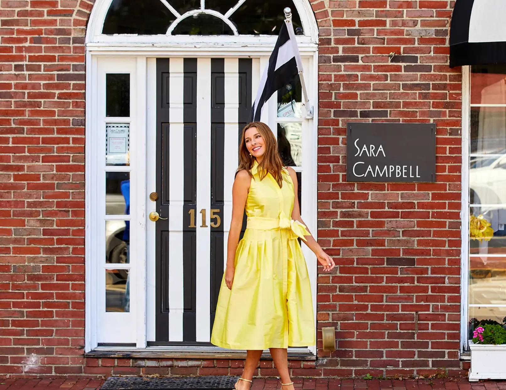 A woman in a yellow belted dress walking past the striped door of the Sara Campbell boutique